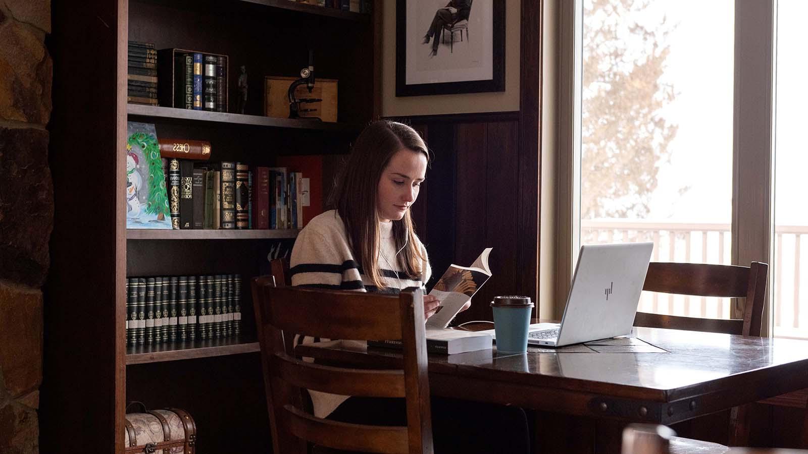 英语学生靠窗阅读，桌上放着咖啡和笔记本电脑