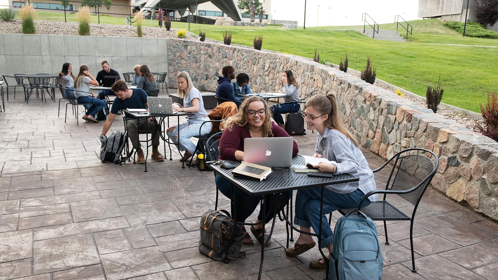Several year-round campus students sitting at tables on a patio studying with laptops open